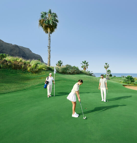 People playing golf on a green course with palm trees and mountains in the background, near the ocean.
