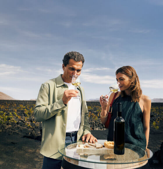 Couple tasting white wine at a glass table with a bottle and appetisers in a volcanic landscape.
