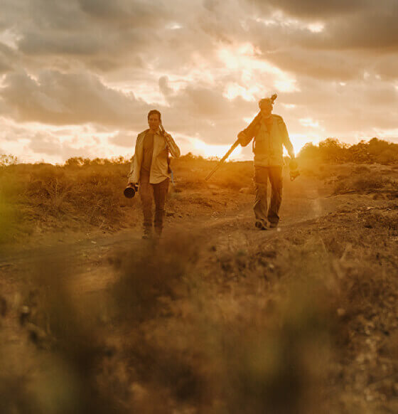 Two people with birdwatching gear walk through an arid, volcanic landscape at dusk.