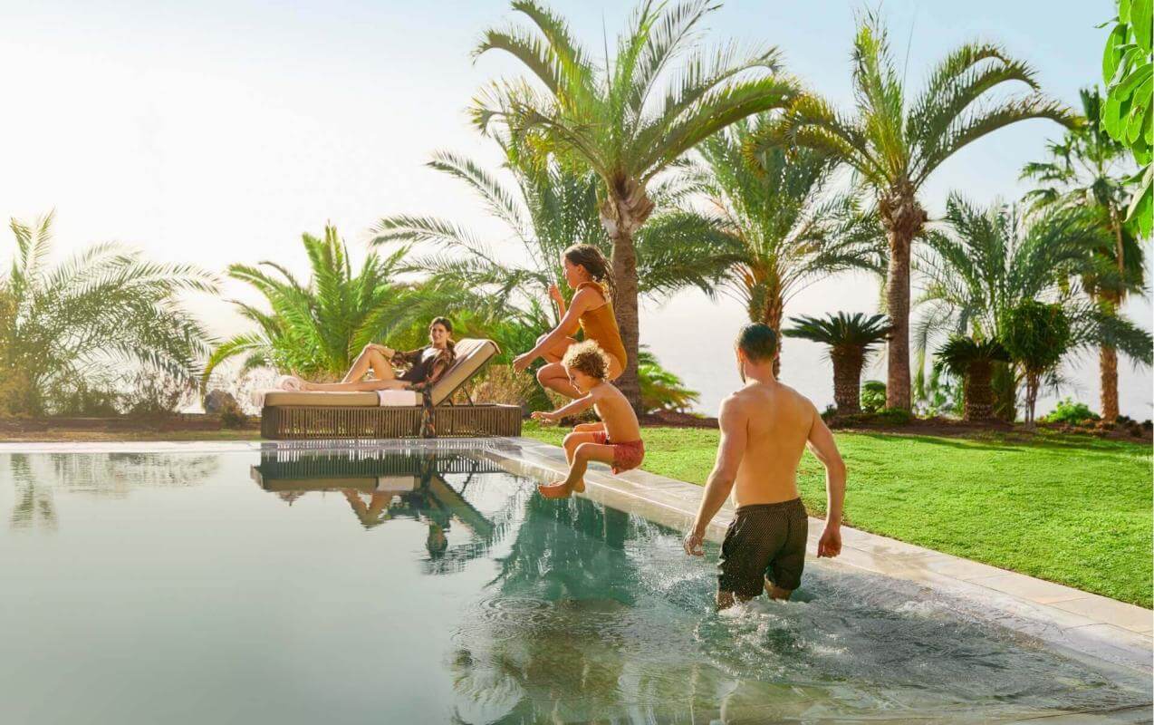 Family enjoying an infinity pool surrounded by palm trees and lawns, in a sunny setting with natural views.