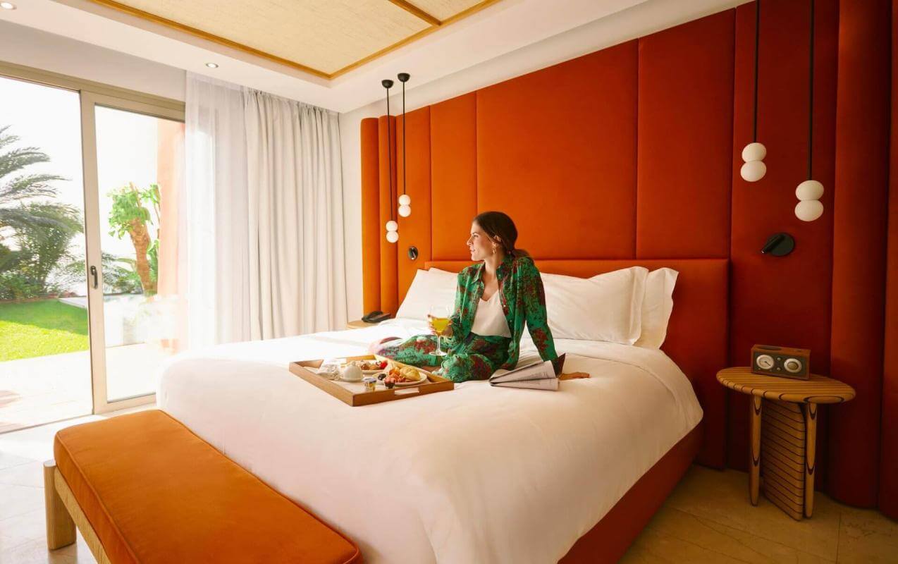 Person sitting on a bed with breakfast served in a bright room, with an orange headboard and garden views.