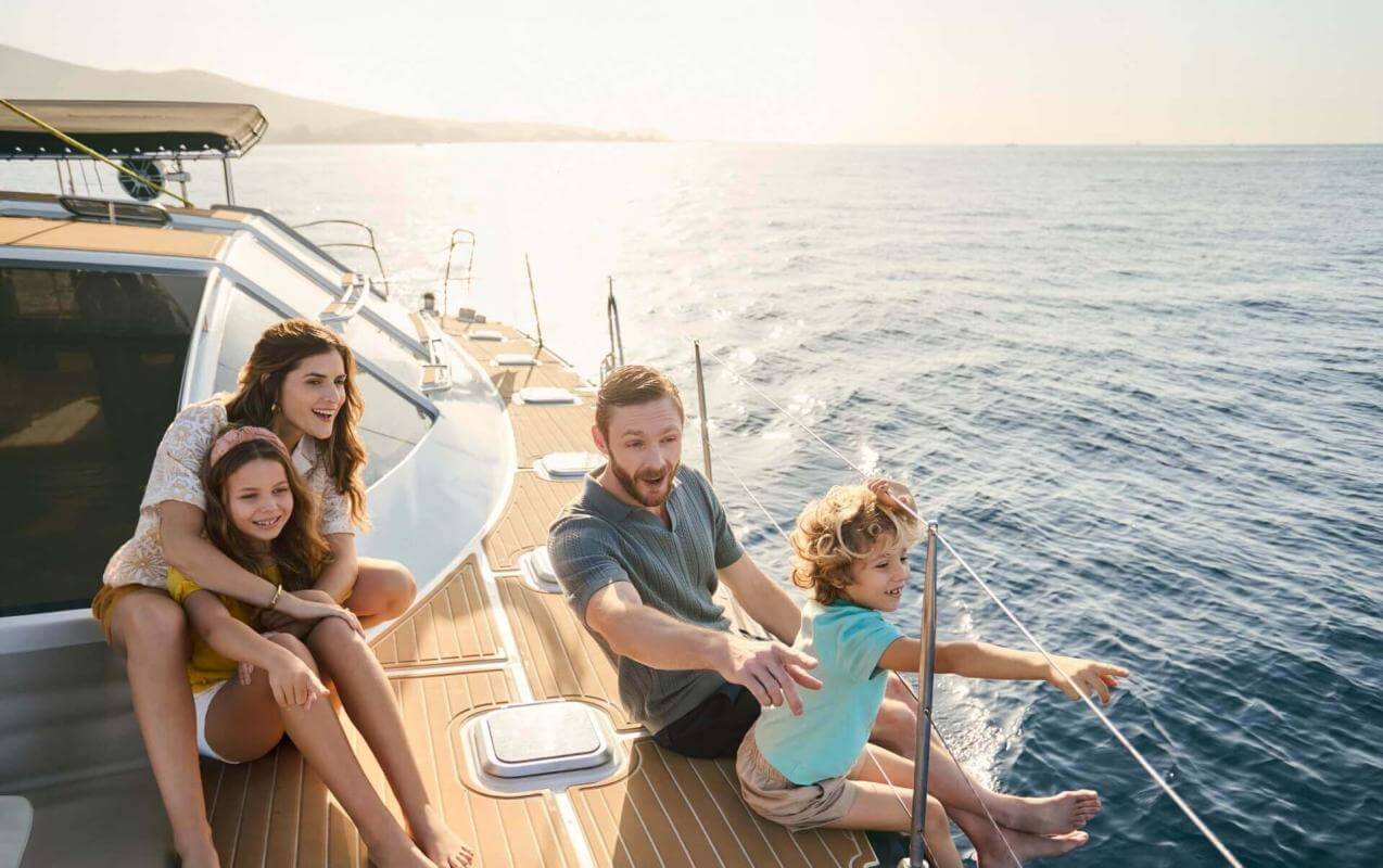 Family relaxing on the deck of a sailing boat at sunset with views of the sea and sunlit coastline.