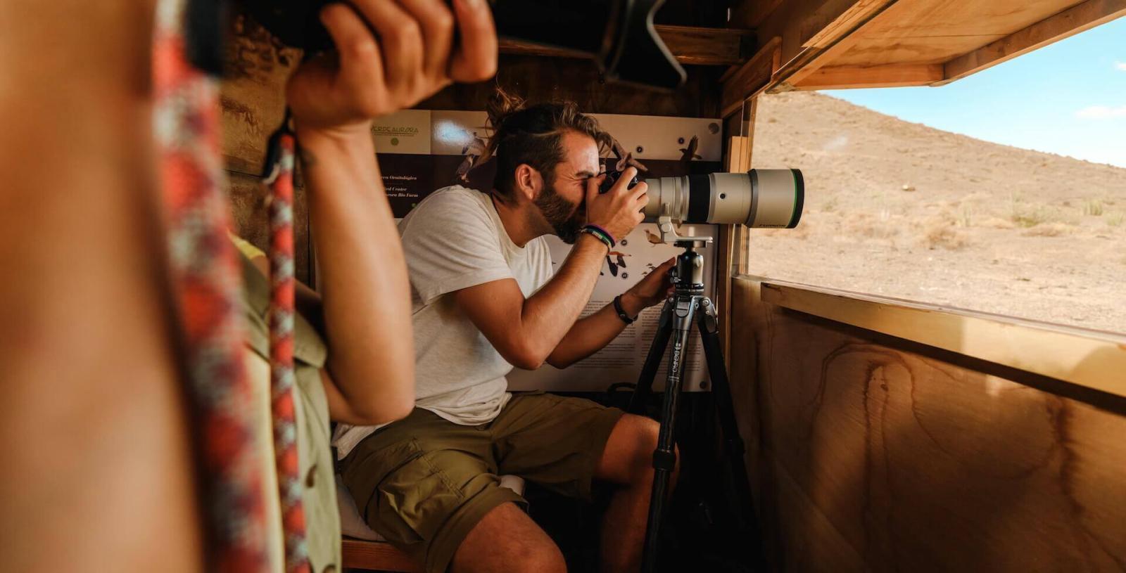 A person photographing fauna with a wide-angle camera from a wooden hideout, focusing on the exterior.