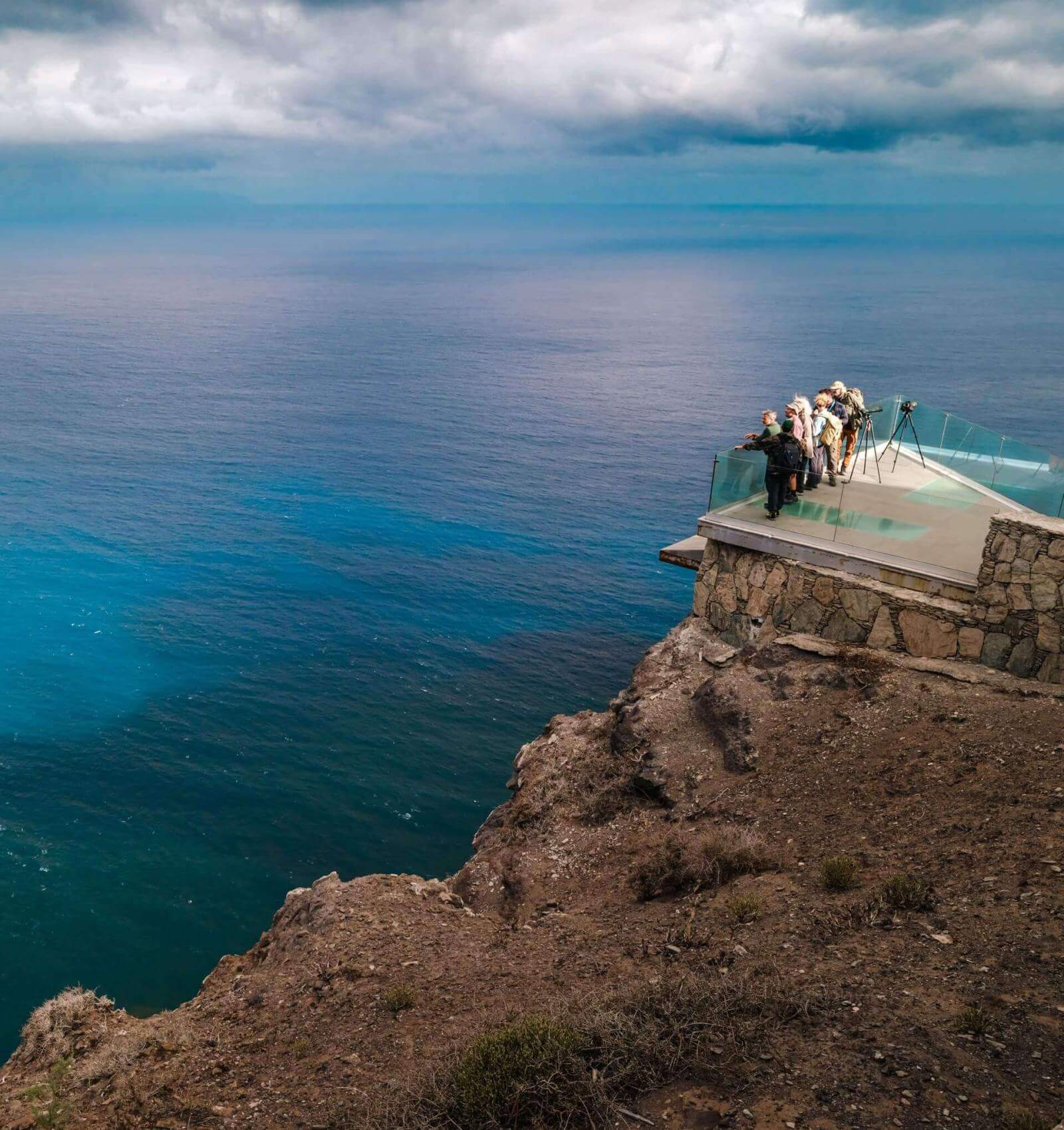 Visitors viewing the Atlantic Ocean from glass viewpoint on cliff under cloudy sky in the Canary Islands.