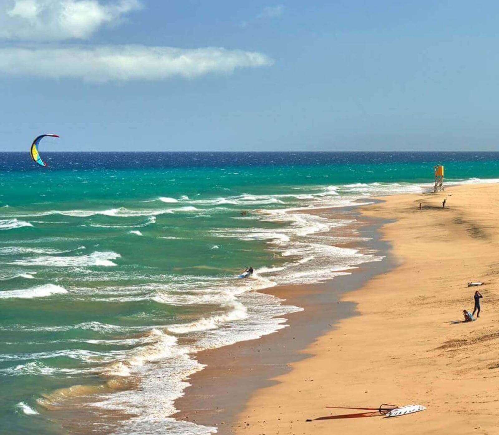 Beach of golden sand with waves and people practising water sports like kitesurfing.