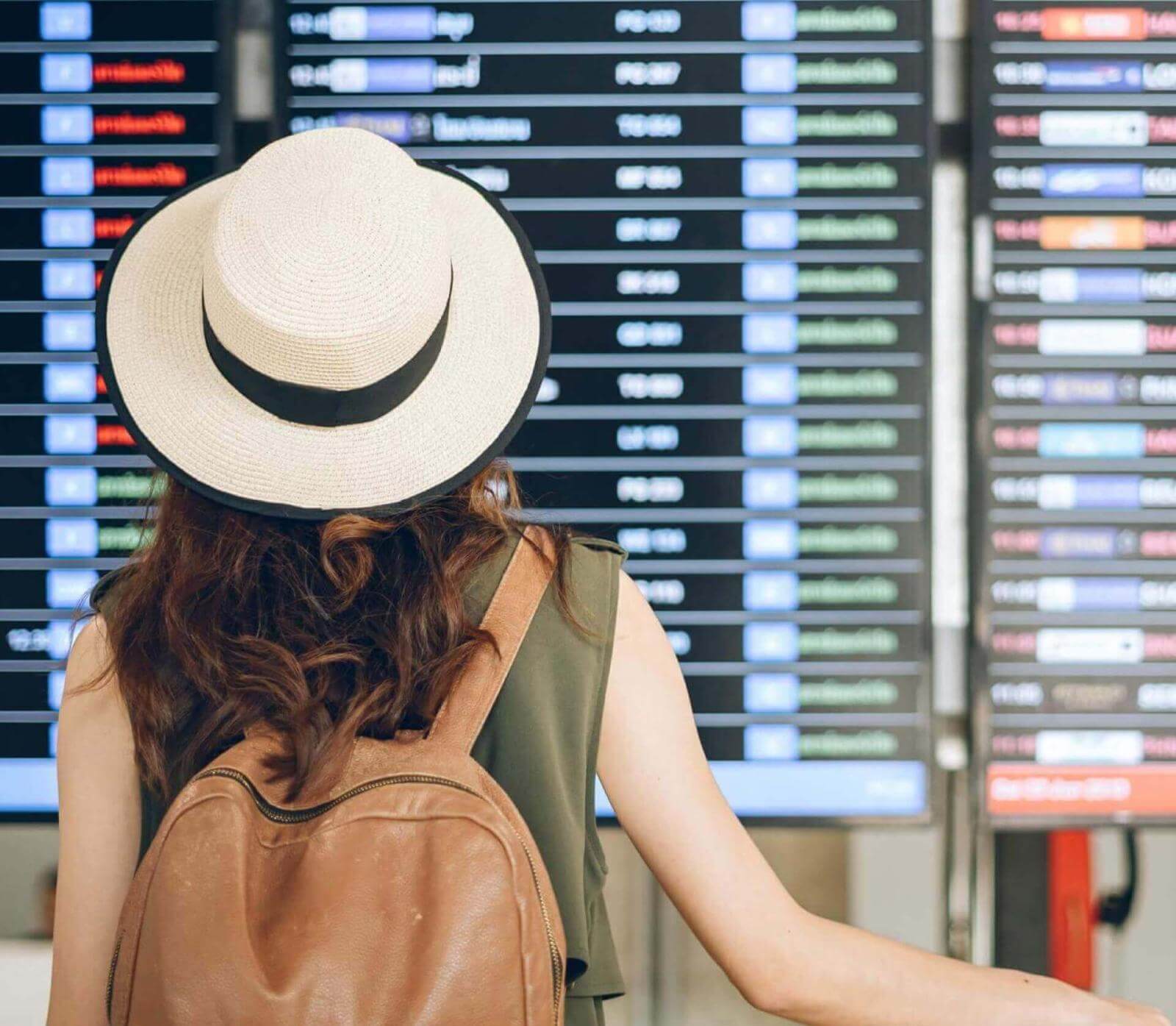 Person with hat and backpack looking at flight information display in an airport.