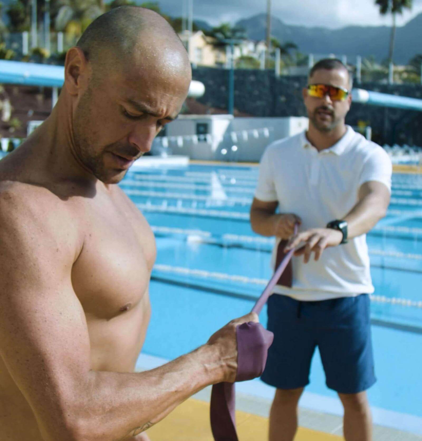 Person performing strength exercises with a resistance band next to an Olympic pool, supported by a trainer.