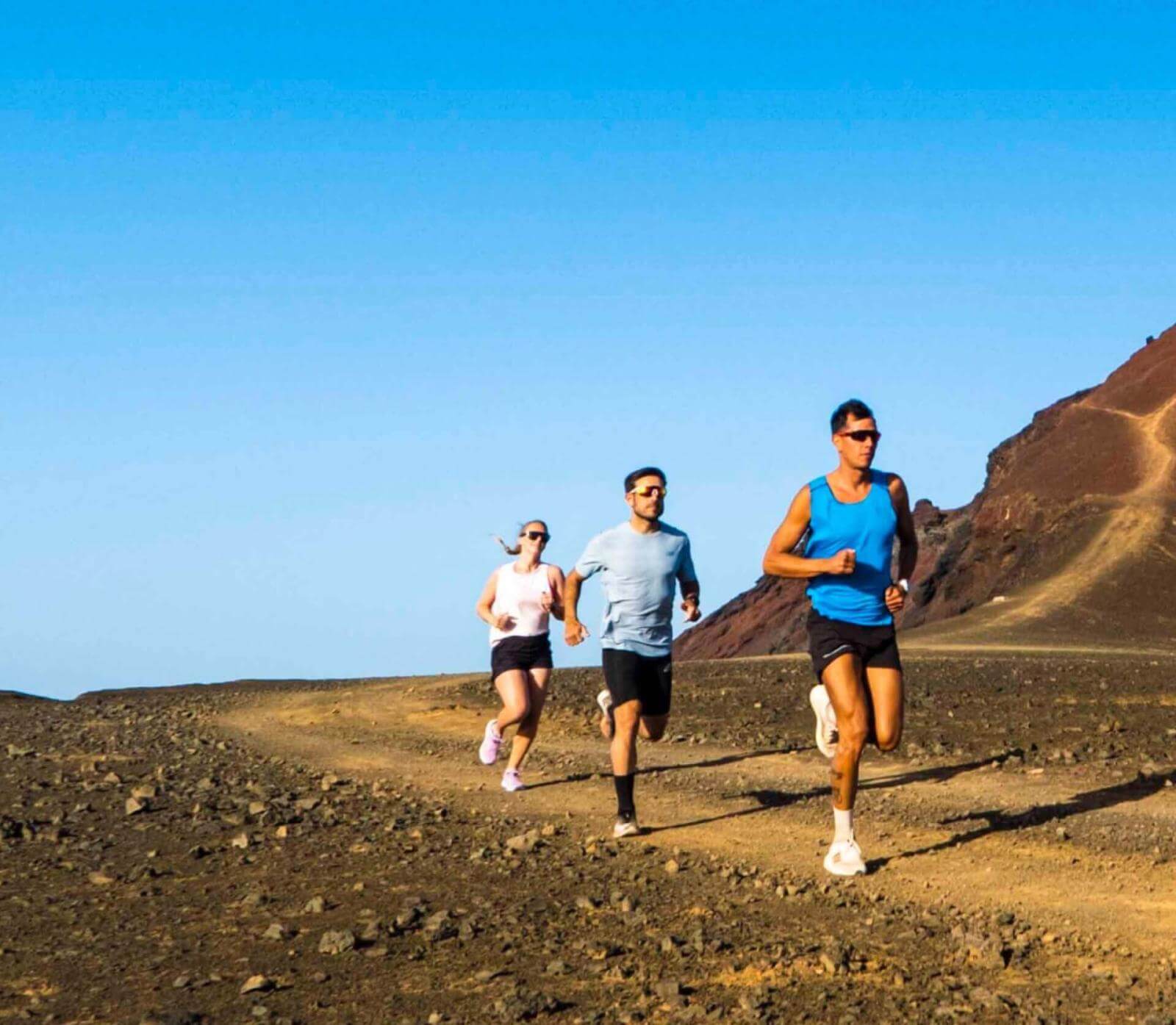 Three athletes training on a wide volcanic terrain trail under a clear sky.
