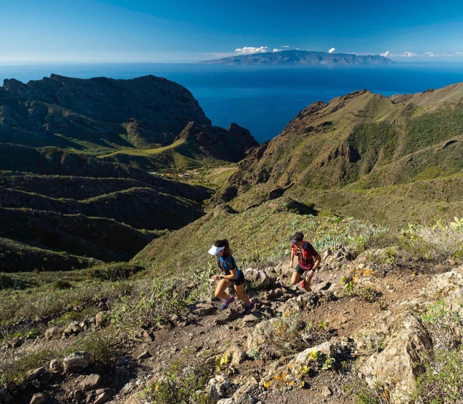 Two people trail running on a mountainous path with ocean views and another island on the horizon.
