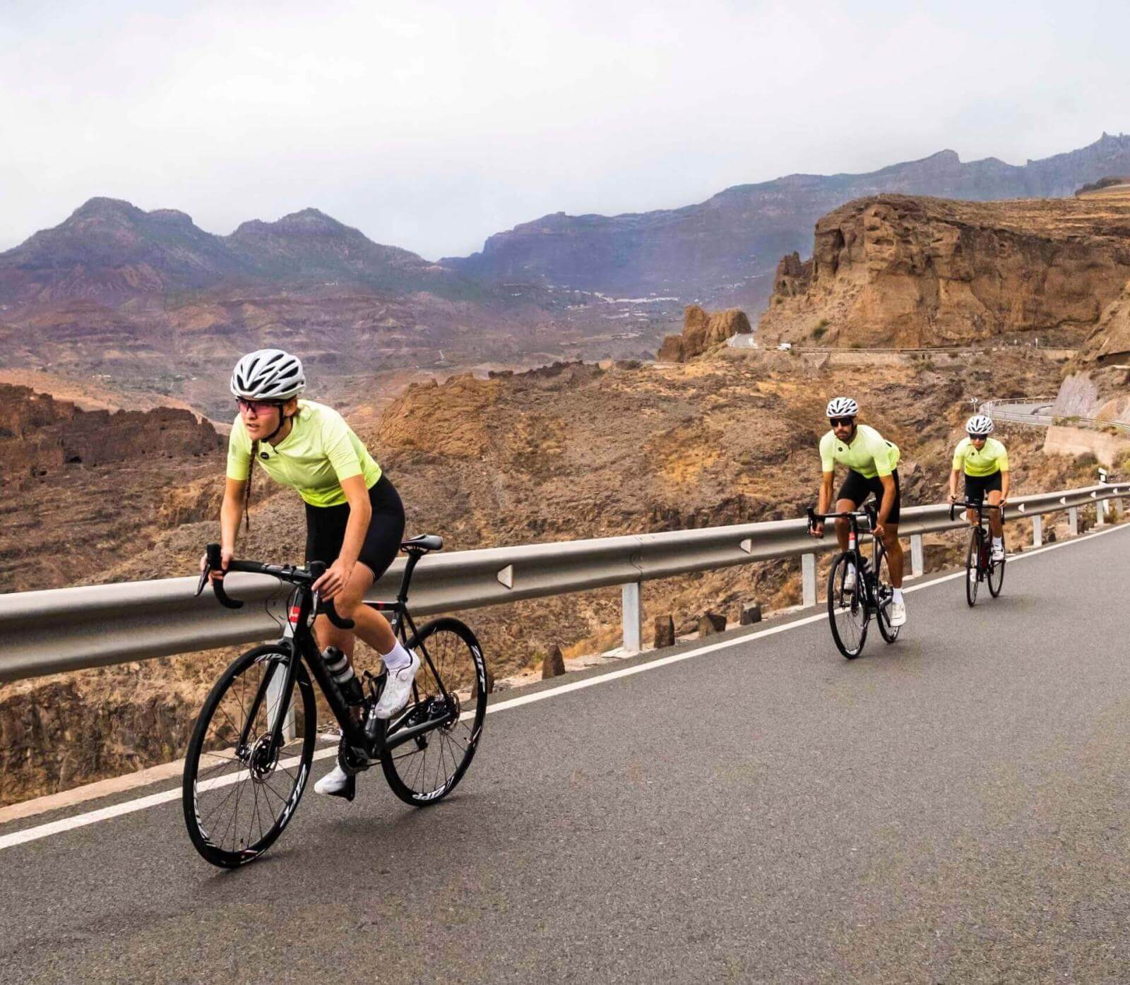 Three cyclists training on an asphalt road surrounded by a mountainous landscape.