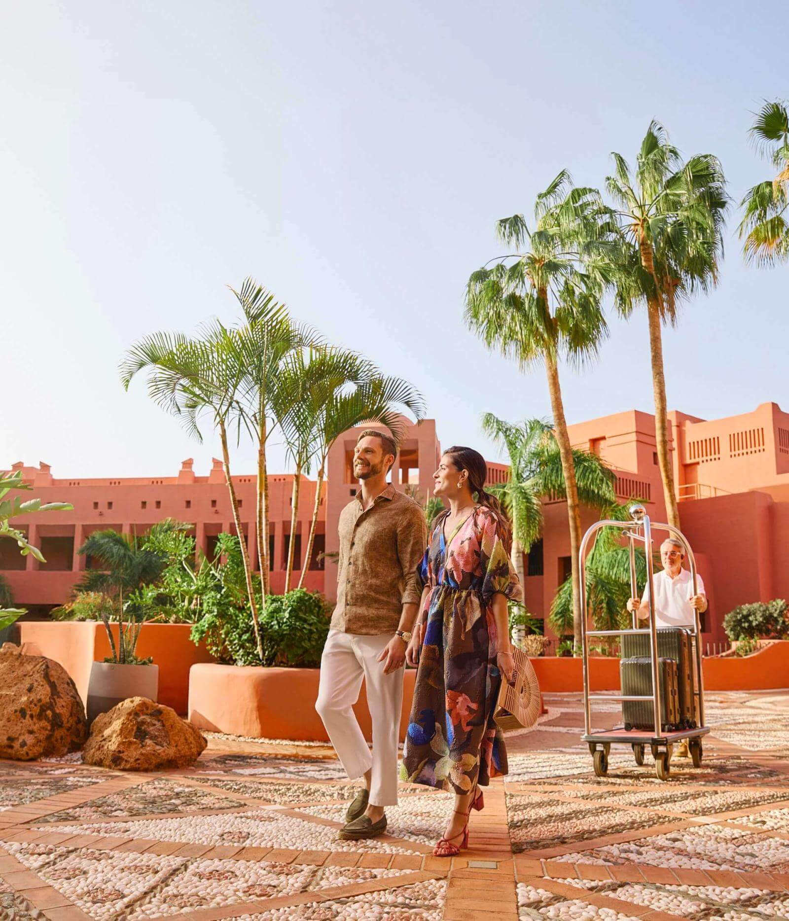 Exterior of a hotel in terracotta tones with palm trees and mosaics; a couple walk while an employee carries luggage.