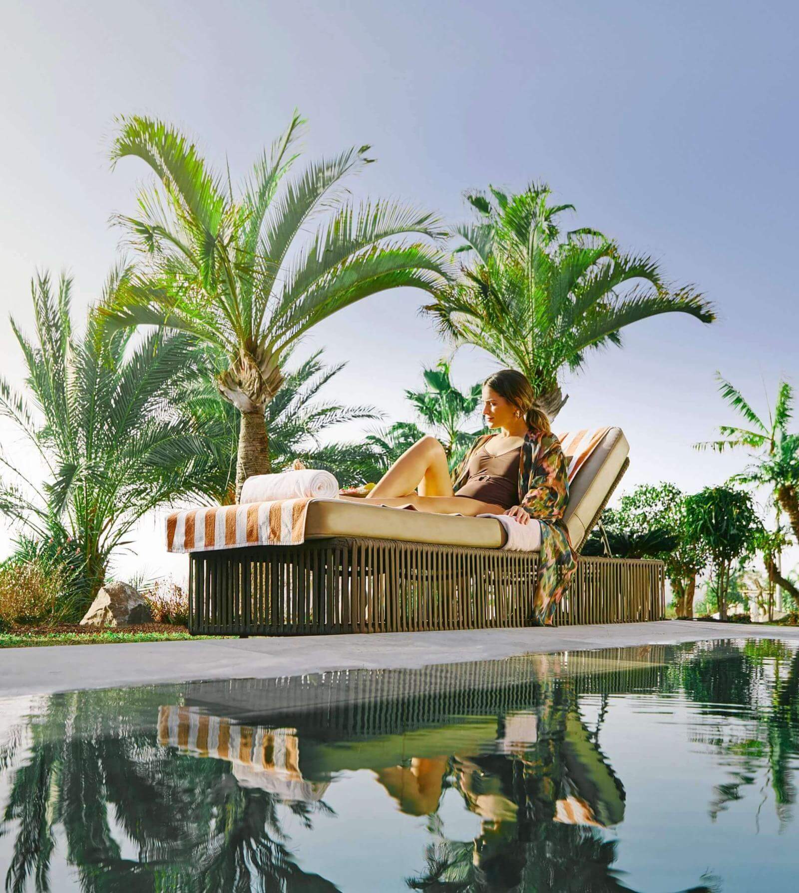 Person relaxing on a sun lounger by a swimming pool surrounded by palm trees, with reflections in the water and clear skies.