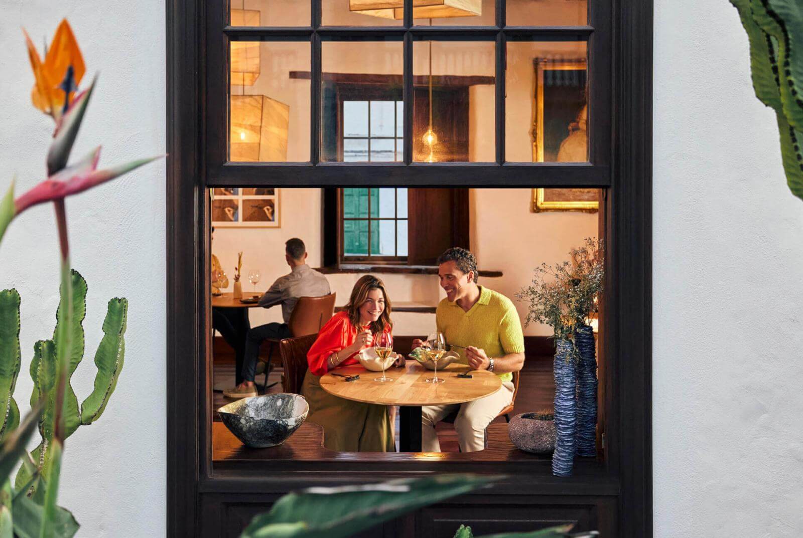 Two people seated at a round table enjoying a meal indoors, seen from a window with plants in the foreground.