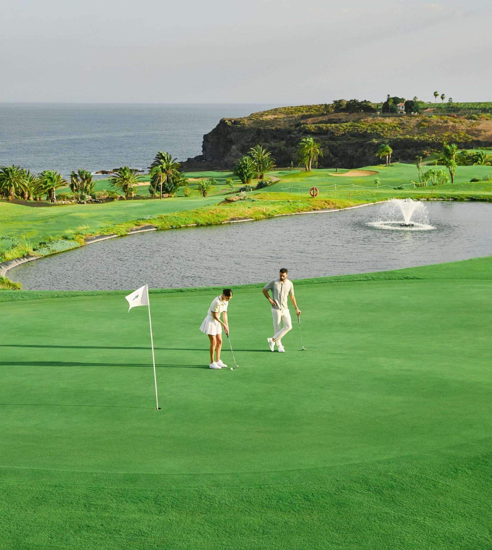 People playing golf on a green next to a lake with a fountain, cliffs, and the sea in the background.