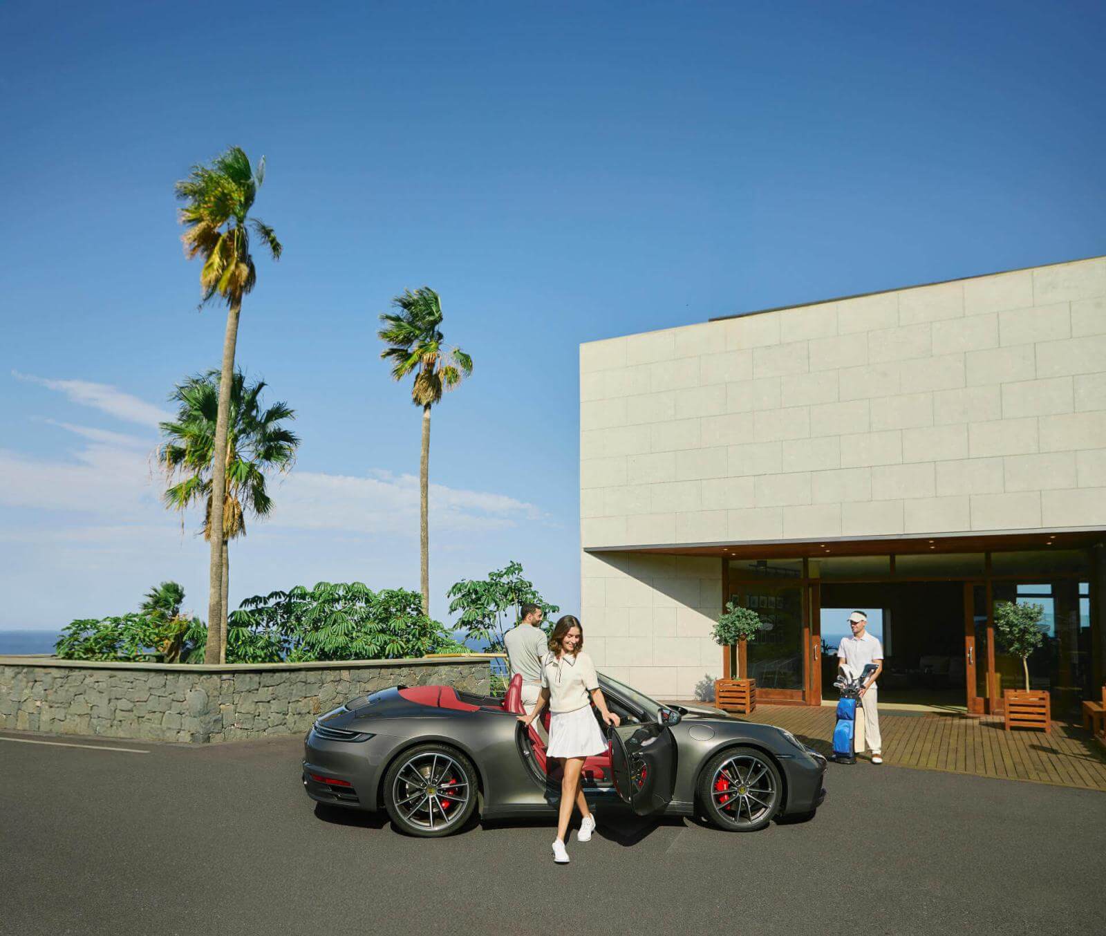 People next to a sports car in front of a modern building with palm trees and a caddie waiting.