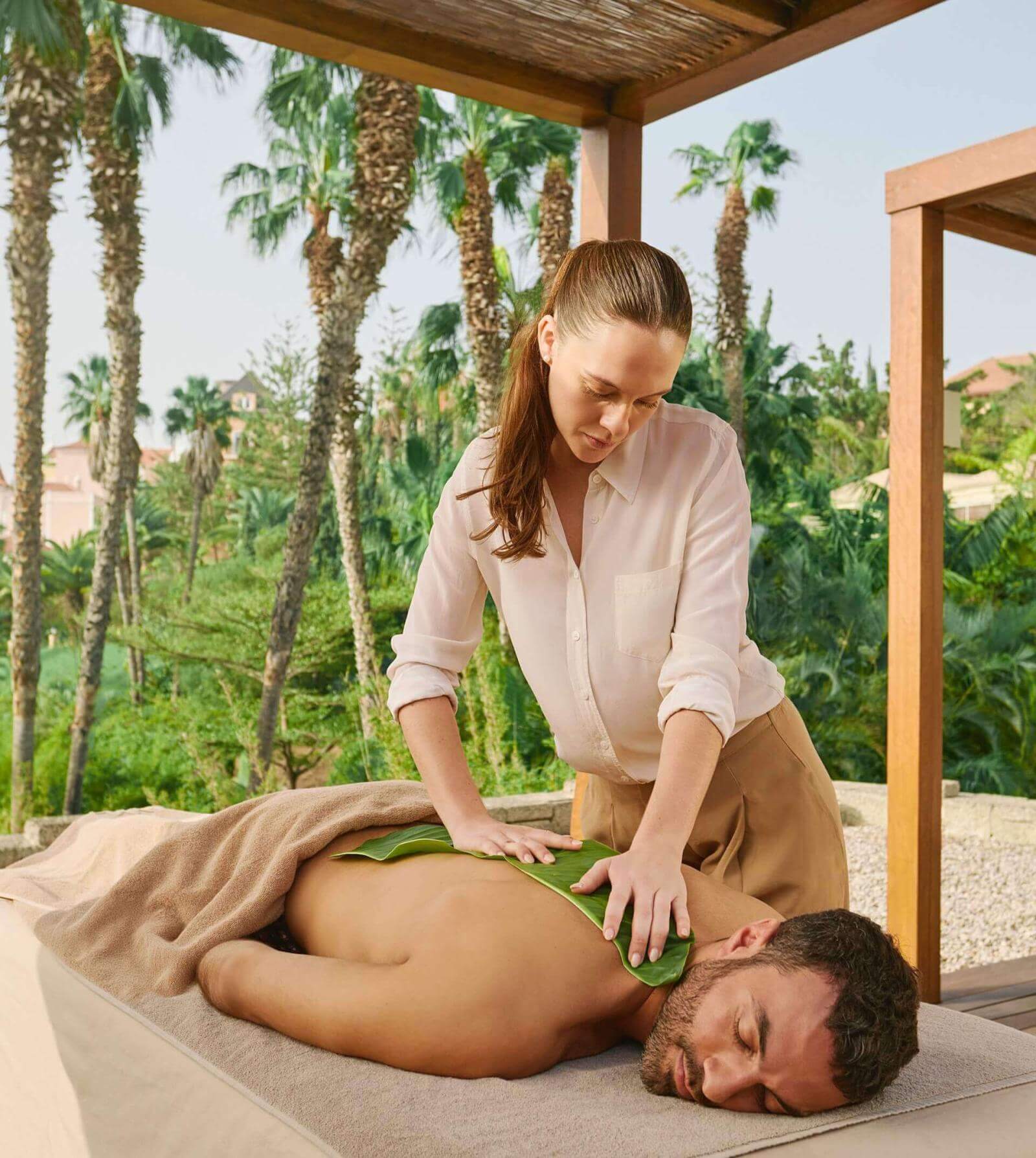 Person enjoying a massage in a hotel garden with palm trees under a pergola. 
