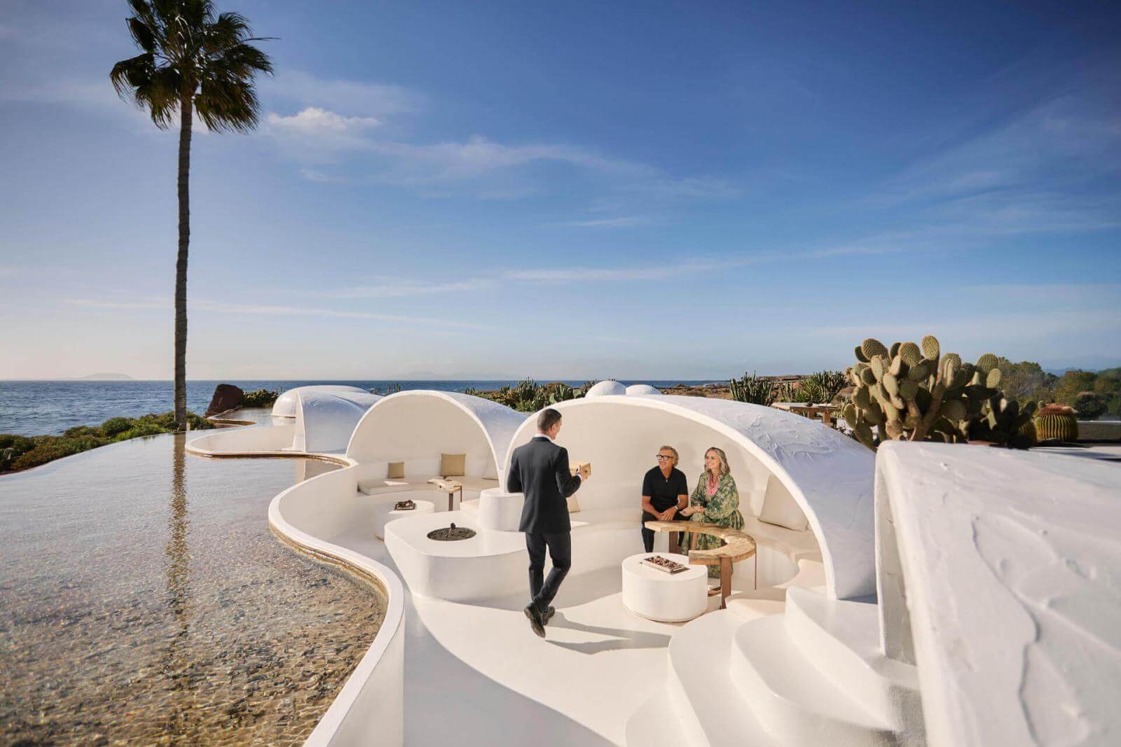 People conversing on a white terrace with organic design next to a pool, with sea views.