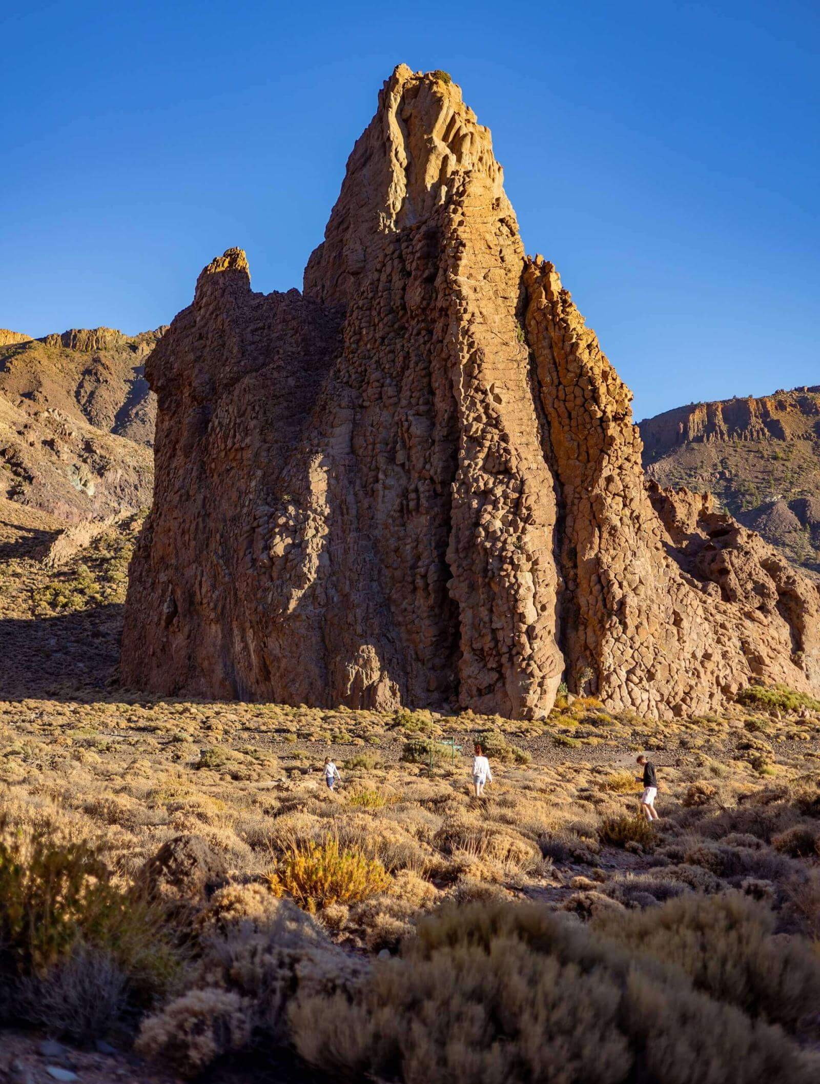 People walking through low scrub in front of a large vertical rock formation in a volcanic landscape.