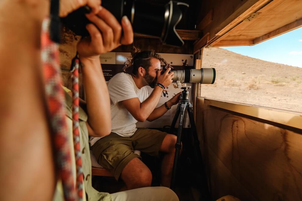 A person photographing fauna with a wide-angle camera from a wooden hideout, focusing on the exterior.