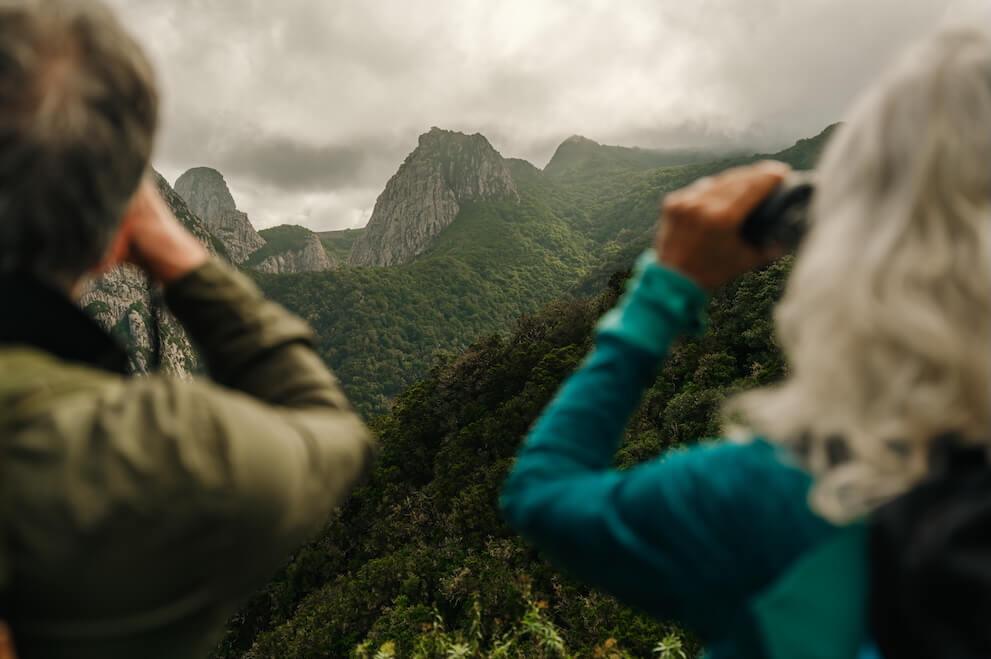 Birdwatchers with binoculars in front of forest-covered mountains under cloudy sky, Canary Islands.