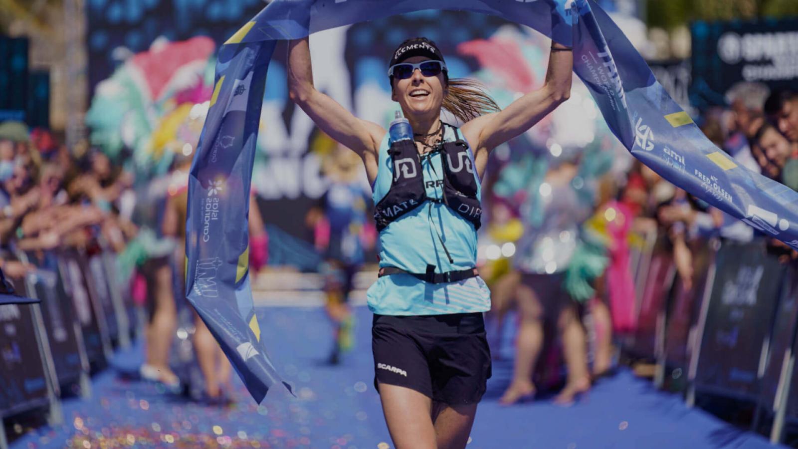Person crossing the finish line at an outdoor sports competition with tape held up and a cheering crowd.