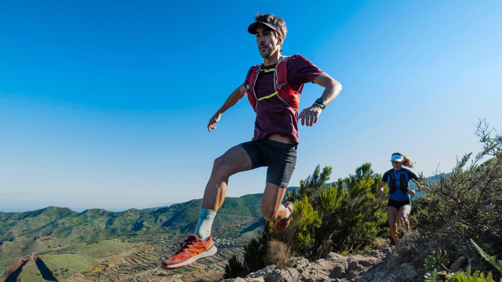People running on a rocky trail in a mountainous natural environment with vegetation and a clear sky.