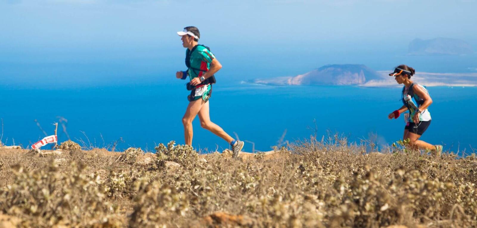 Pair of athletes training on a mountainous trail with a volcanic landscape and the sea in the background.