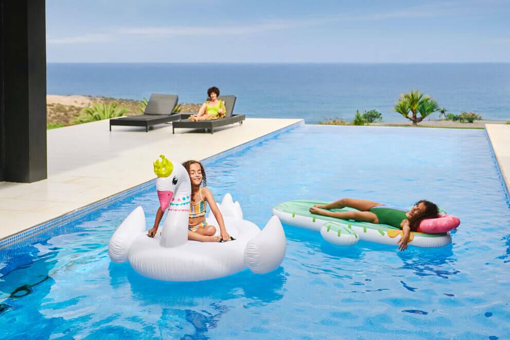 Children playing in a swimming pool with floats while an adult relaxes in the background, with the ocean visible on the horizon.