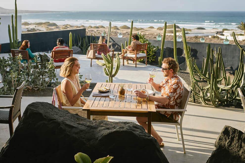 Two people sitting on an outdoor terrace enjoying food and drink, with cacti and ocean views.