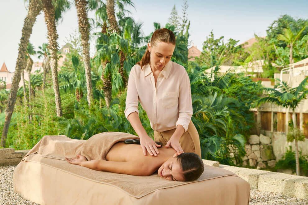 Therapist performing a relaxing massage outdoors on a massage table, surrounded by palm trees and tropical vegetation.