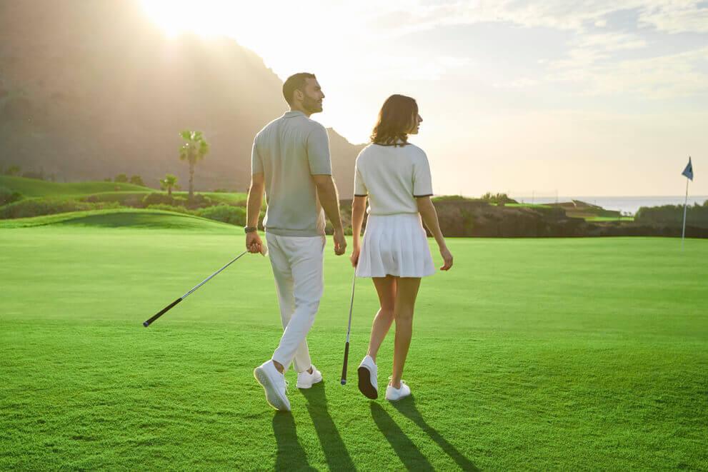 Two people walking on the green of a golf course at sunset, with a volcanic landscape.