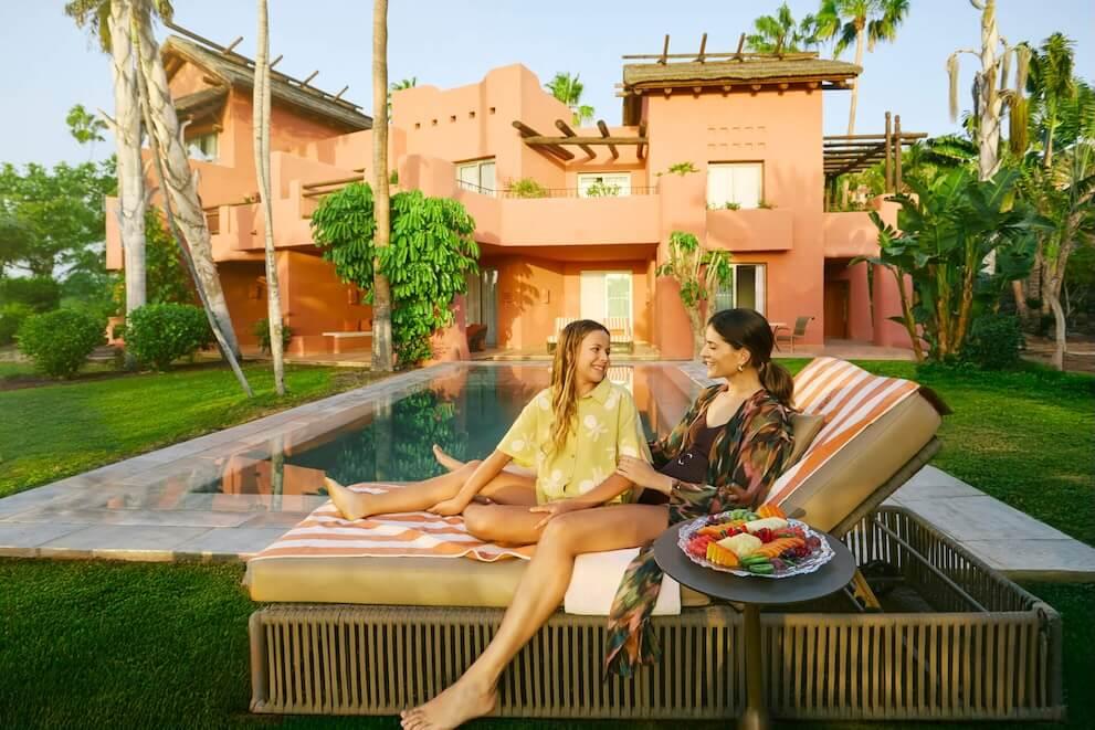 Two people relaxing on a sun lounger by the pool, with a fruit platter, in front of a building surrounded by tropical vegetation.