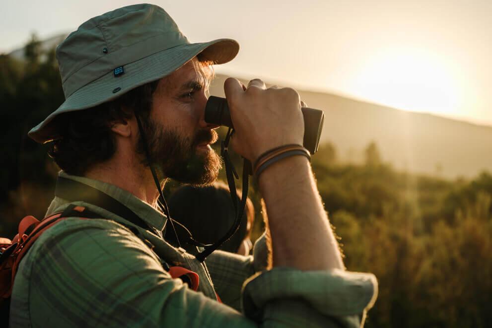 Close-up of a person wearing a hiking hat observing the landscape through binoculars at sunset.