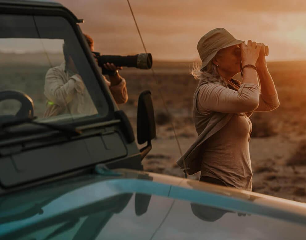 A person birdwatching with binoculars in an arid landscape at sunset next to a 4x4 vehicle.
