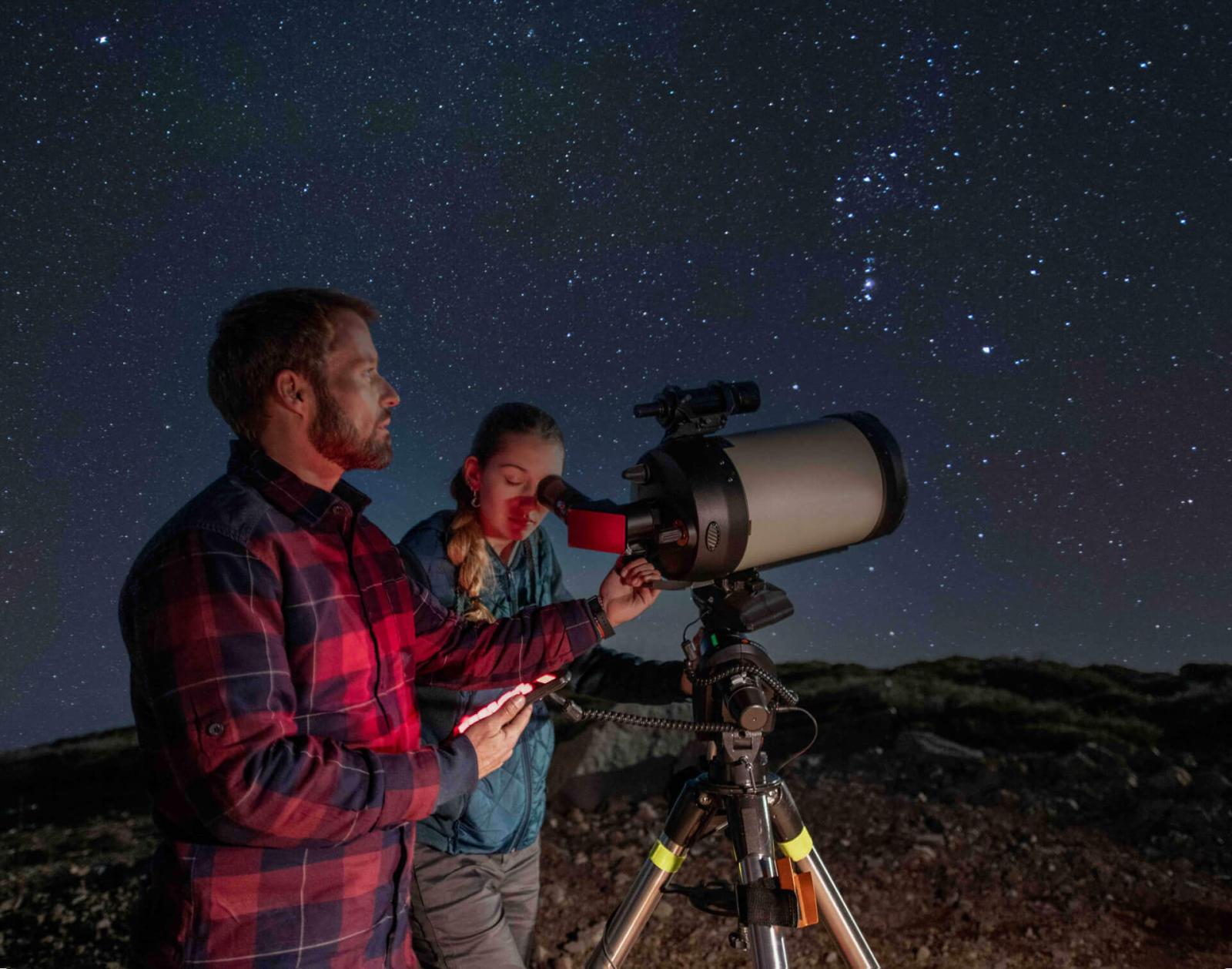 Two people using a telescope under a starry night sky in a rocky landscape.