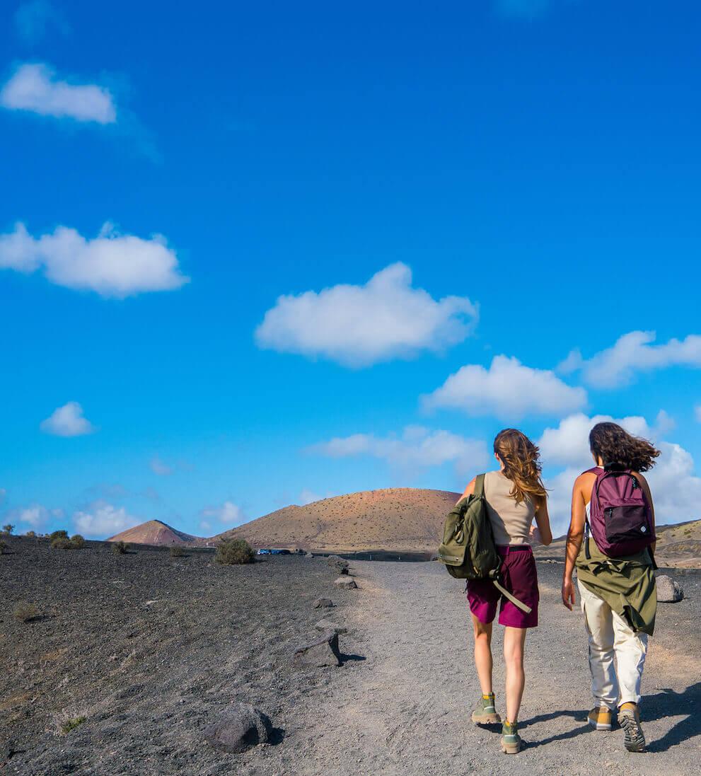 Two people walking a volcanic trail in Timanfaya National Park under a blue sky.