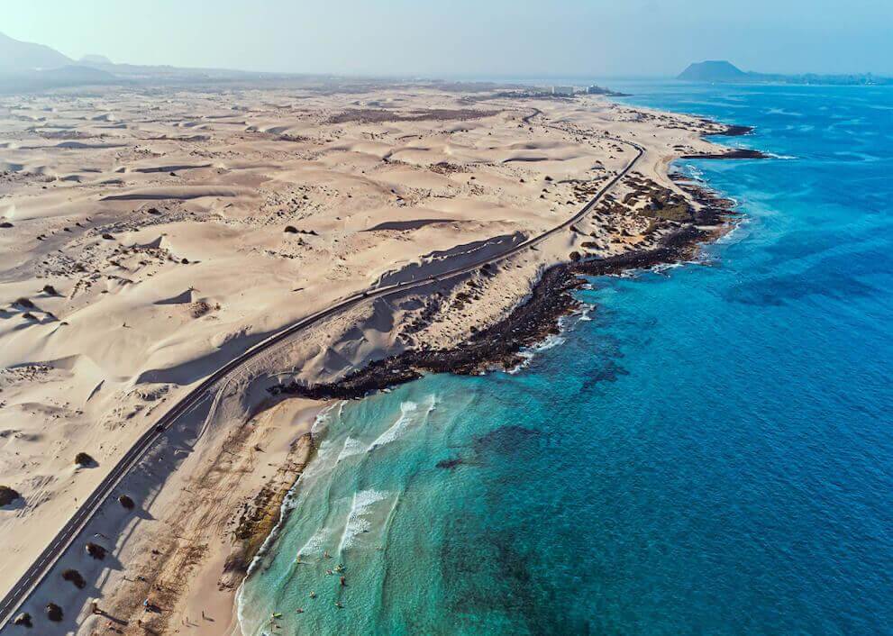 Aerial view of Fuerteventura’s Corralejo Dunes: vast golden sands by a turquoise sea and a volcanic coastline.