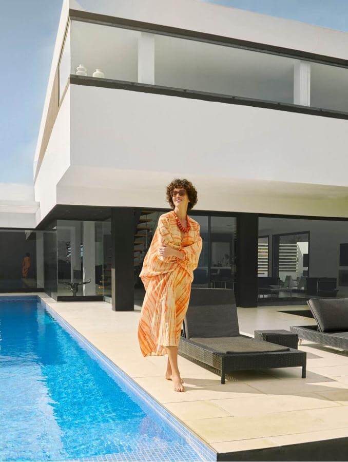 Person beside a private pool in front of a modern villa with minimalist lines, with sun loungers and clear skies.