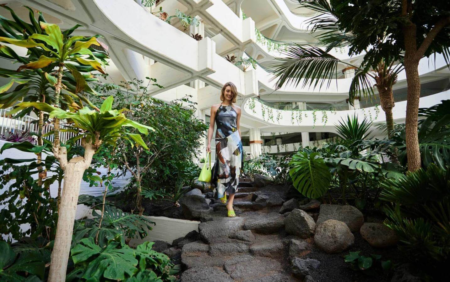 Person walking along a path surrounded by tropical vegetation in an interior with white architecture and organic design.