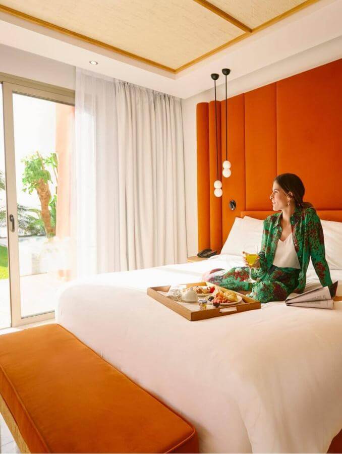 Person sitting on a bed with breakfast served in a bright room, with an orange headboard and garden views.