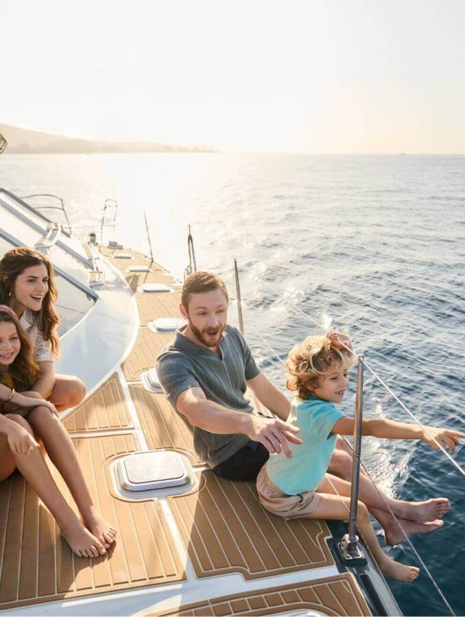 Family relaxing on the deck of a sailing boat at sunset with views of the sea and sunlit coastline.