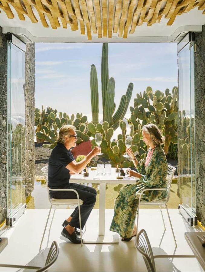 Two people enjoying food in front of the door of a bright terrace surrounded by cacti, in a modern, welcoming space.